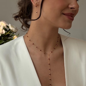 Puede incluir: Un collar de oro y pendientes a juego con pequeñas cuentas de color rojo oscuro. El collar tiene un diseño en forma de Y, con las cuentas espaciadas uniformemente a lo largo de la delicada cadena de oro. Los pendientes son largos y colgantes, con una disposición similar de cuentas.