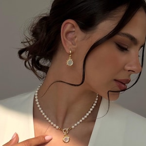 Puede incluir: Un conjunto de joyas doradas con un collar de perlas y pendientes a juego. El collar tiene un colgante en forma de sol con un centro de perla. Los pendientes son pequeños aros con un dije en forma de sol.