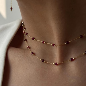 Op de afbeelding: Een delicate gouden ketting met twee strengen, elk versierd met kleine, ronde, dieprode kralen. Een bijpassende oorbel met één rode kraal is zichtbaar. De sieraden zijn geplaatst tegen een neutrale achtergrond.