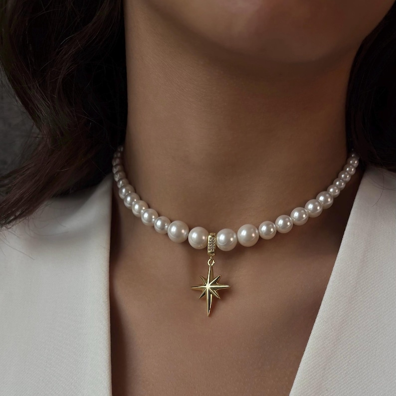 May include: A pearl necklace with a gold star pendant. The necklace features a strand of white pearls of varying sizes. The gold pendant is a star shape with eight points. The necklace is worn on a person wearing a white blazer.