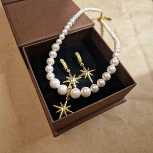 May include: A pearl necklace and matching earrings with gold starburst pendants are displayed in a brown gift box. The earrings have a small hoop design with a starburst pendant. The necklace and earrings are set against a black velvet interior.