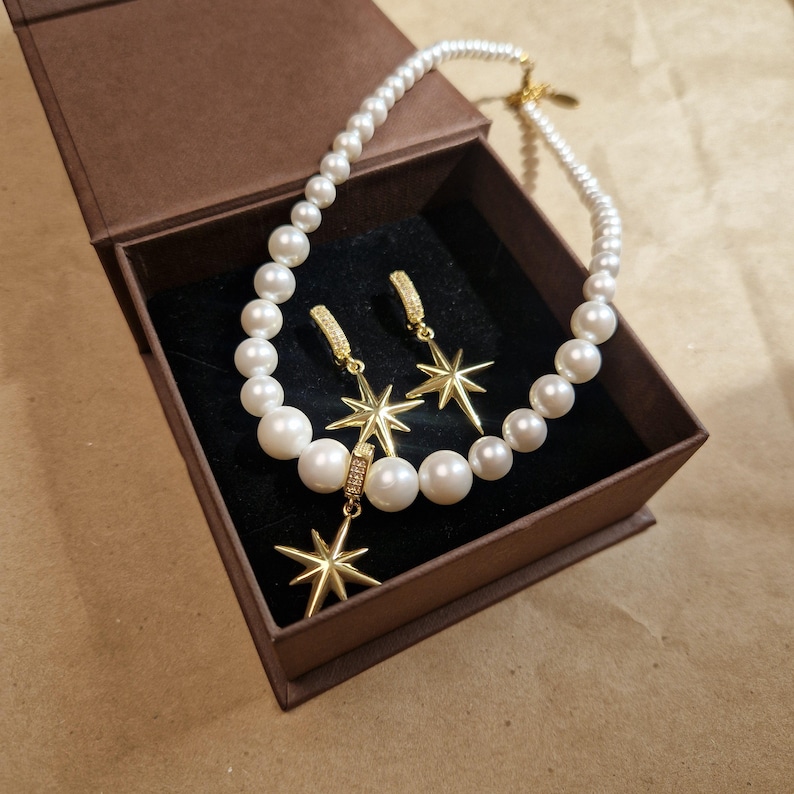 May include: A pearl necklace and matching earrings with gold starburst pendants are displayed in a brown gift box. The earrings have a small hoop design with a starburst pendant. The necklace and earrings are set against a black velvet interior.