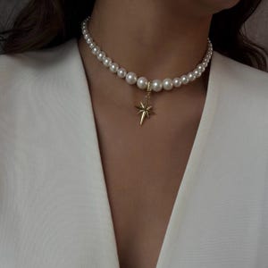 May include: A pearl necklace with a gold star pendant. The necklace features multiple white pearls and a gold starburst charm. The star pendant hangs from a small gold loop. The necklace is worn against a white blazer.