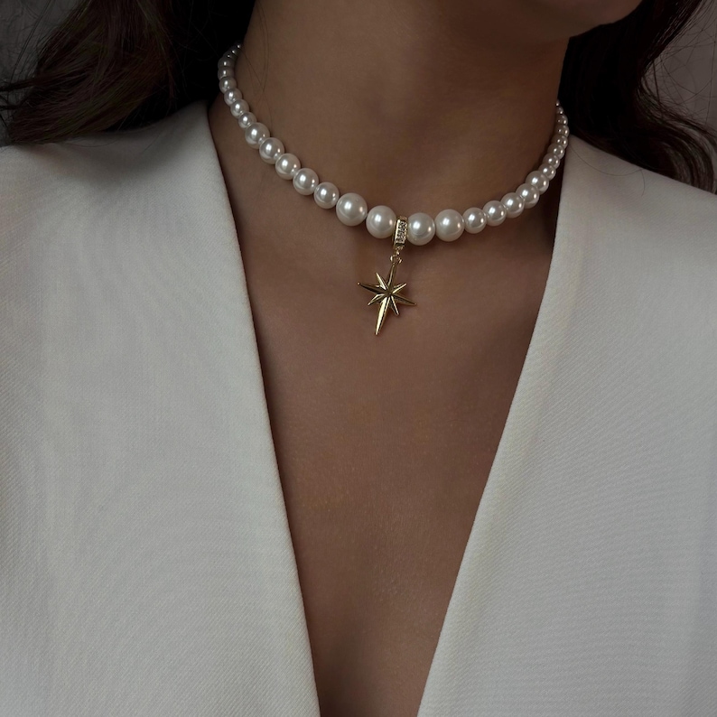 May include: A pearl necklace with a gold star pendant. The necklace features multiple white pearls and a gold starburst charm. The star pendant hangs from a small gold loop. The necklace is worn against a white blazer.