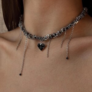 K&ouml;nnte beinhalten: Eine silberne Choker-Kette mit einem schwarzen Herz-Anh&auml;nger und baumelnden Ketten. Die Kette besteht aus mehreren miteinander verwobenen Ketten und hat einen zarten, kantigen Look.