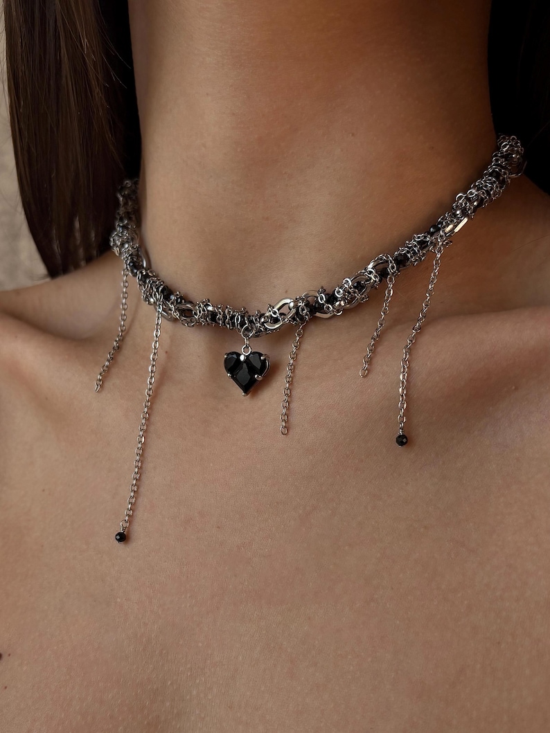 K&ouml;nnte beinhalten: Eine silberne Choker-Kette mit einem schwarzen Herz-Anh&auml;nger und baumelnden Ketten. Die Kette besteht aus mehreren miteinander verwobenen Ketten und hat einen zarten, kantigen Look.