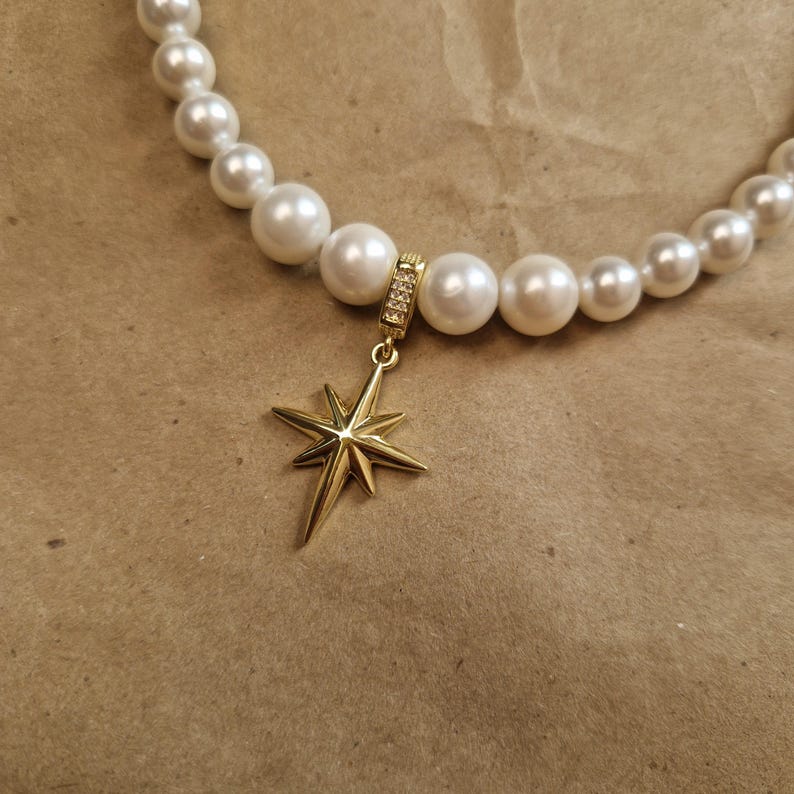 May include: A pearl necklace with a gold star pendant. The necklace features large, round, white pearls. The star pendant is gold-colored and has a multi-pointed design. The pendant's bail is adorned with small, clear crystals.