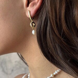 May include: Gold hoop earrings with a small pearl dangling from a black and gold charm. The earrings are worn by a person with long brown hair. The person is also wearing a pearl necklace.