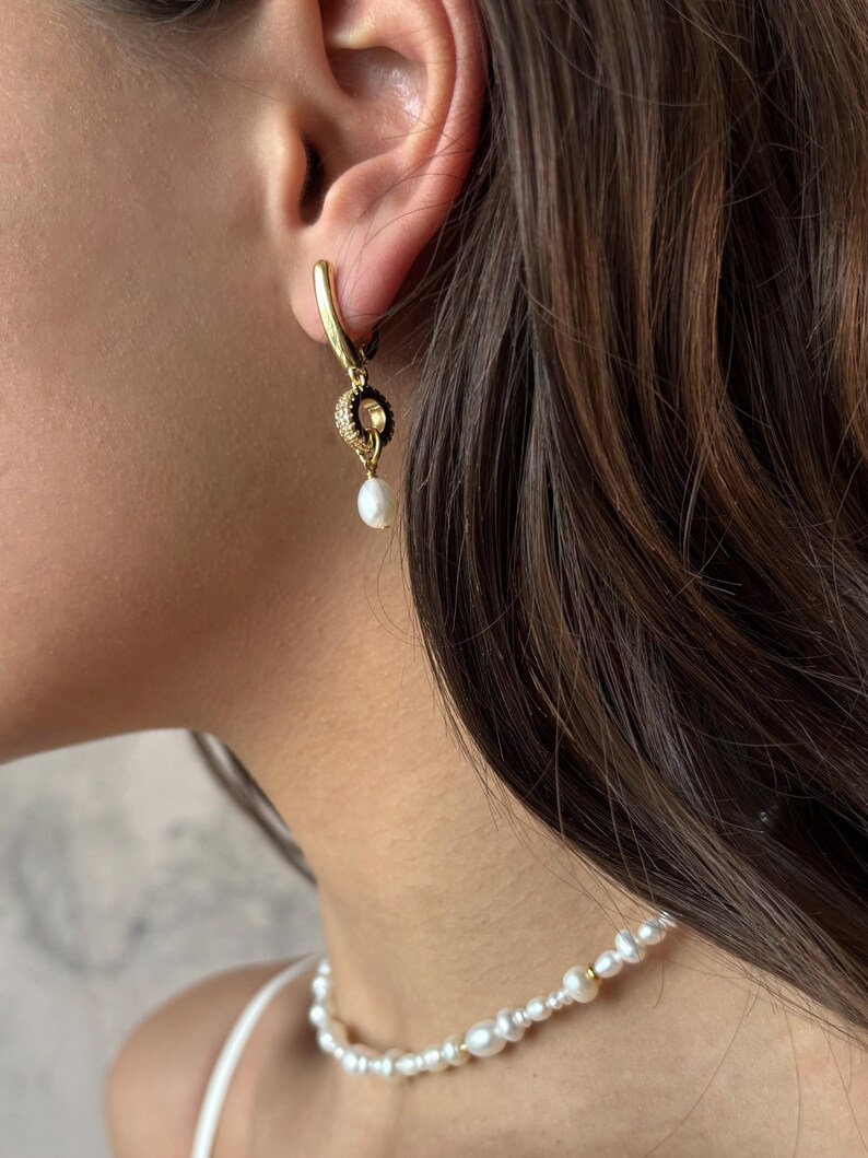 May include: Gold hoop earrings with a small pearl dangling from a black and gold charm. The earrings are worn by a person with long brown hair. The person is also wearing a pearl necklace.