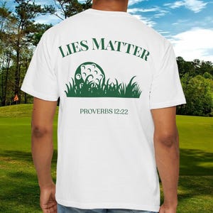 May include: White t-shirt with a green graphic on the back. The graphic features a golf ball in grass with the words "LIES MATTER" above and "PROVERBS 12:22" below. The shirt is being worn on a golf course.