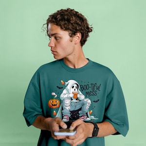 以下が含まれることがあります： ハロウィーンをテーマにしたグラフィックが特徴の、ティールグリーンのオーバーサイズTシャツ。白いフード付きマントを着たゴーストキャラクターがマグカップを持ち、「BOO-tiful mess」の文字が描かれています。ジャック・オー・ランタンとキャンディーコーンも含まれています。