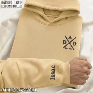 以下が含まれることがあります： 上部に「EMBROIDERED DAD EST. HOODIE」の文字が入ったベージュ色のパーカー。 パーカーには「EST. 2024」と袖に「Issac」の名前が入った「DAD」ロゴが入っています。 下部には「FATHER'S DAY SPECIAL」の文字が入っています。