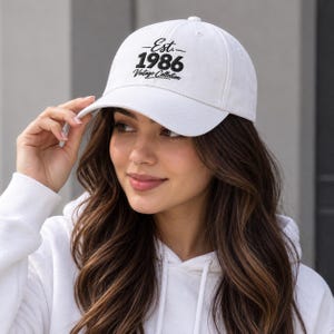 May include: A white baseball cap featuring black embroidered text that reads Est. 1986 Vintage Collection. The cap has a structured crown, a curved brim, and a button on top. The design is centred on the front panel of the white fabric hat.