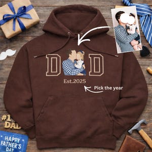 May include: A brown hooded sweatshirt with a front pocket and a drawstring hood. The front displays the word "DAD" in a block letter design, with a custom illustration of a father and child. The text "Est.2025" is below the illustration, with the words "Pick the year" nearby.