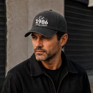 May include: A charcoal grey baseball cap featuring white embroidered text that reads Est. 1986 Vintage Collection. The cap has a structured crown, a curved brim, and a washed fabric finish, providing a classic look with clear, legible lettering.