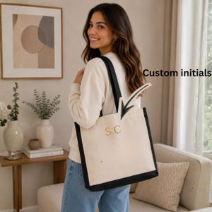 Puede incluir: Un bolso tote de lona color crema con bordes y asas negras. La parte frontal del bolso presenta las iniciales S.C. bordadas en dorado y una flecha negra dibujada a mano que apunta hacia el texto Custom initials.