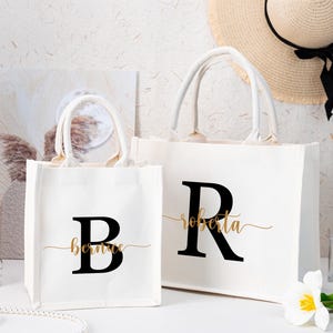 Può includere: Due borse tote in tela bianca con scritte nere e dorate. La borsa più piccola ha una grande "B" nera con "Bernice" in corsivo dorato sotto. La borsa più grande ha una grande "R" nera con "Roberta" in corsivo dorato.