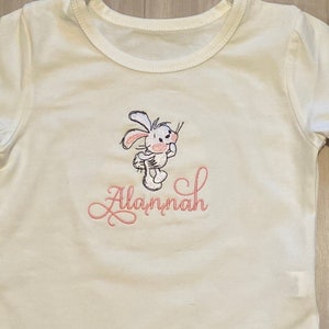 May include: White baby bodysuit with a pink embroidered bunny and the name "Alannah" in pink cursive lettering.