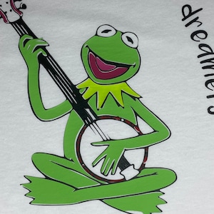 May include: A green frog playing a banjo with the text "The lovers, the dreamers and me" on a white t-shirt.
