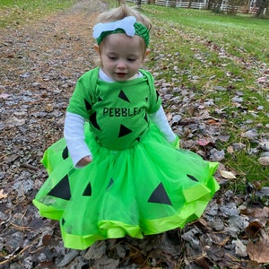May include: A young child dressed as Pebbles Flintstone for Halloween. The child is wearing a green and black tutu with a green shirt that says "Pebbles" on it. The child is also wearing a white bone headband.
