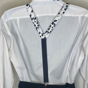 May include: Black suspenders with a white pattern of Jack Skellington's face from the Nightmare Before Christmas movie. The suspenders are worn over a white button-down shirt and black pants.