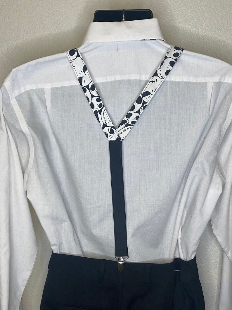 May include: Black suspenders with a white pattern of Jack Skellington's face from the Nightmare Before Christmas movie. The suspenders are worn over a white button-down shirt and black pants.