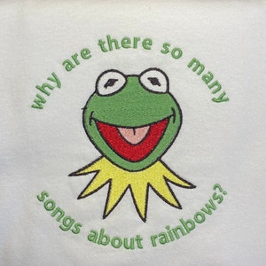 May include: A white sweatshirt with a green frog embroidered on the front. The frog is wearing a yellow collar and has a big smile. The text around the frog reads "Why are there so many songs about rainbows?"