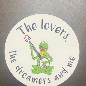 May include: A white round coaster with a green frog playing a banjo. The text "The lovers, the dreamers and me" is written around the frog.