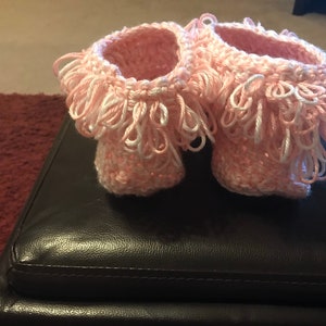 May include: A pair of pink crocheted baby booties with white fringe trim.