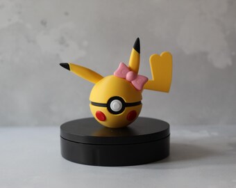 May include: A yellow Pikachu figure with a pink bow, black-tipped ears, and a red and white design on its cheeks. The figure is on a black circular base, set against a neutral background.