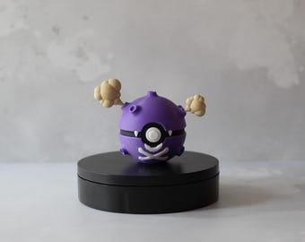 May include: A purple, spherical figurine with two beige, cloud-like appendages. The figurine has a black stripe, a white eye, and a skull and crossbones design. It sits on a black, circular base.
