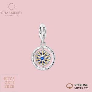 Puede incluir: Un charm de plata con un diseño de brújula. El charm presenta una piedra azul central rodeada de piedras doradas y transparentes, engastada en un anillo de plata. La marca "CHARMLEFY" está en la parte superior, con "BRACELET CHARMS" debajo. Las palabras "STERLING SILVER 925" están en la parte inferior.