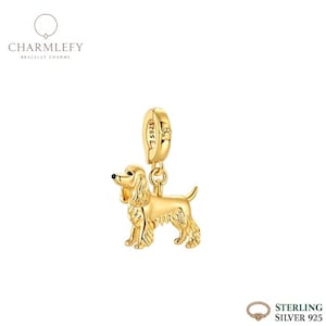 Może przedstawiać: Złoty charms w kształcie psa, prawdopodobnie Cocker Spaniel, zawieszony na pętli. Charms jest szczegółowy z teksturowanym futrem i posiada małą pętlę do mocowania. Słowo "CHARMLEFY" znajduje się w lewym górnym rogu.