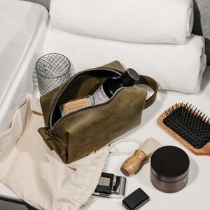 May include: An olive green leather toiletry bag with a zipper, handle, and various grooming products. The bag contains a bottle, soap, and other items. A wooden hairbrush, shaving brush, and a jar of cream are also visible.