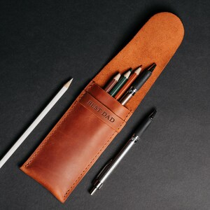May include: A brown leather pencil case with the embossed text "BEST DAD". The case holds several pencils and pens, including a white pencil and a silver pen. The case is open, revealing the contents against a black background.