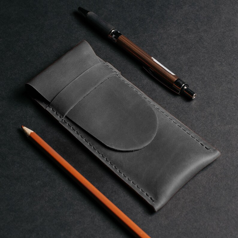 May include: A dark gray leather pen case with a flap closure, a wooden pen, and an orange pencil on a black surface. The pen has a silver tip and black ink. The pencil is sharpened and ready for use. The pen case is stitched with a contrasting thread.