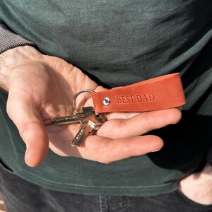 May include: A brown leather keychain with the text "BEST DAD" embossed on it. The keychain is attached to a set of keys.