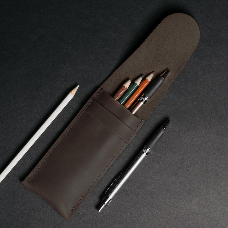 May include: A brown leather pencil case with a flap, holding several colored pencils and pens. A white pencil and a black pen are placed beside the case on a dark surface. The case is designed to hold writing instruments.