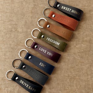 May include: A set of nine leather keychains with different phrases embossed in gold lettering. The keychains are in various colors, including black, brown, red, green, and purple. The phrases include "Sweet Home", "Best Dad", "Sunshine", "Freedom", "Love You", "EDC", "W&R 2024", and "Drive Safe".