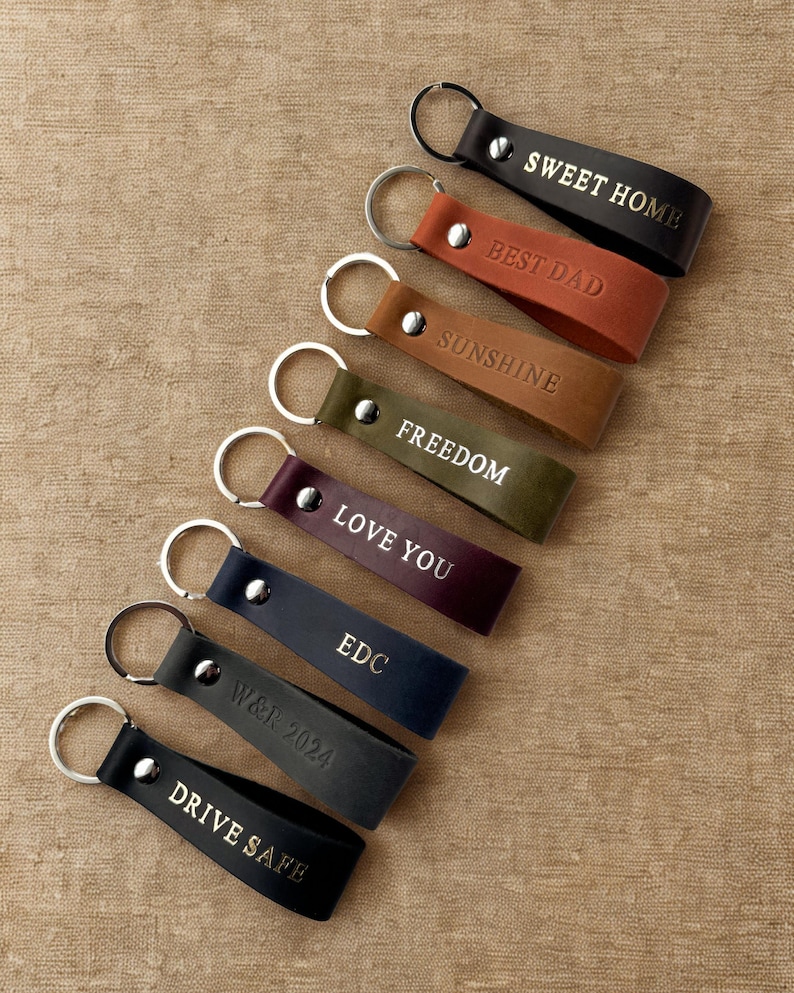 May include: A set of nine leather keychains with different phrases embossed in gold lettering. The keychains are in various colors, including black, brown, red, green, and purple. The phrases include "Sweet Home", "Best Dad", "Sunshine", "Freedom", "Love You", "EDC", "W&R 2024", and "Drive Safe".