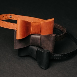 Pode incluir: Três gravatas borboleta de couro em tons de castanho e preto são empilhadas sobre uma superfície escura. A gravata borboleta superior é castanho claro, a do meio é castanho escuro e a inferior é preta. Cada gravata borboleta é presa a uma tira de couro a condizer.