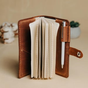 Könnte beinhalten: Ein brauner Notizbuchumschlag aus Leder mit sichtbaren Nähten, der einen Stapel karierter Papierseiten enthält. Ein weißer Stift steckt in einer Lederschlaufe auf der rechten Seite, und ein kleiner Lederriemen mit silbernem Druckknopf sichert den Umschlag.