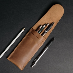 May include: A brown leather pencil case with a flap, holding several pencils and pens. A white pencil and a silver pen are next to the case. The case and writing utensils are on a black surface.
