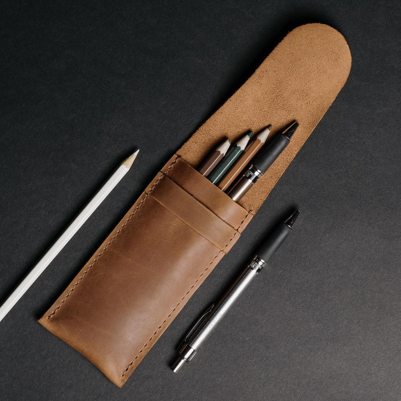 May include: A brown leather pencil case with a flap, holding several pencils and pens. A white pencil and a silver pen are next to the case. The case and writing utensils are on a black surface.