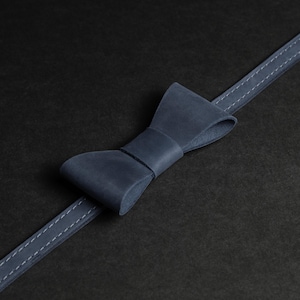 May include: A navy blue leather bow tie with a matching strap, stitched with white thread. The bow tie is centered on a black background, creating a minimalist and elegant aesthetic. The leather appears smooth and well-crafted.