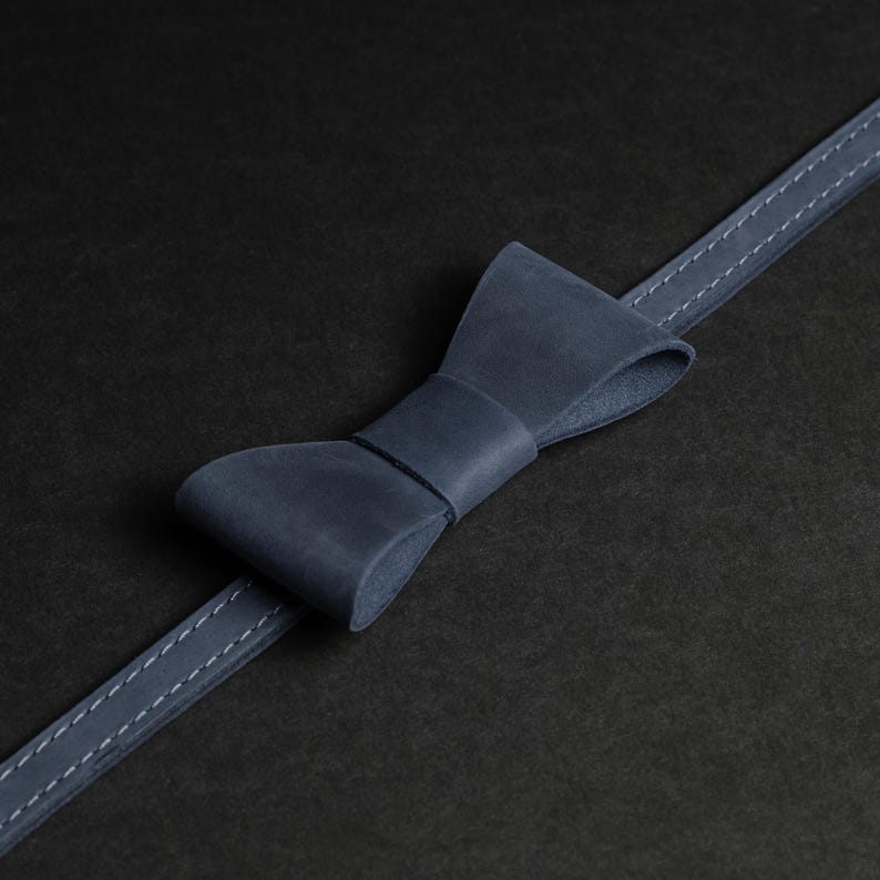 May include: A navy blue leather bow tie with a matching strap, stitched with white thread. The bow tie is centered on a black background, creating a minimalist and elegant aesthetic. The leather appears smooth and well-crafted.