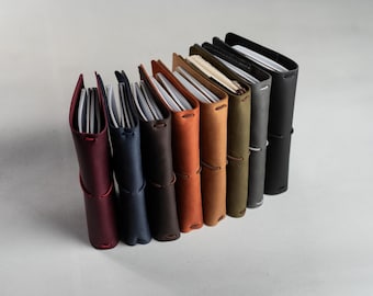 Könnte beinhalten: Eine Sammlung von acht vertikal aufgestellten Notizbüchern mit Ledereinband in einer Reihe, in verschiedenen Farben wie Bordeaux, Marineblau, Dunkelbraun, Hellbraun, Olivgrün, Grau und Schwarz, jeweils mit einem dünnen Gummiband verschlossen.