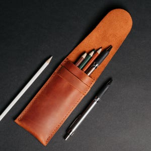 May include: A brown leather pencil case with a flap, holding several colored pencils and pens. A white pencil and a silver pen are placed next to the case on a black surface. The case is made of a smooth, textured leather.