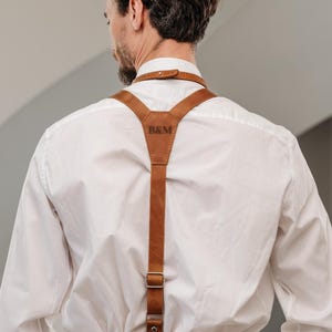 Puede incluir: Tirantes de cuero marrón con las iniciales "B&M" grabadas en la parte trasera. Los tirantes están sujetos a una camisa blanca y pantalones beige. Los tirantes tienen herrajes metálicos y están diseñados para vestimenta formal.