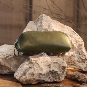 May include: Olive green leather pencil case with a zipper closure and a small loop. The case is resting on a large, textured rock. The stitching is visible along the top seam. The background is a blurred, neutral tone.